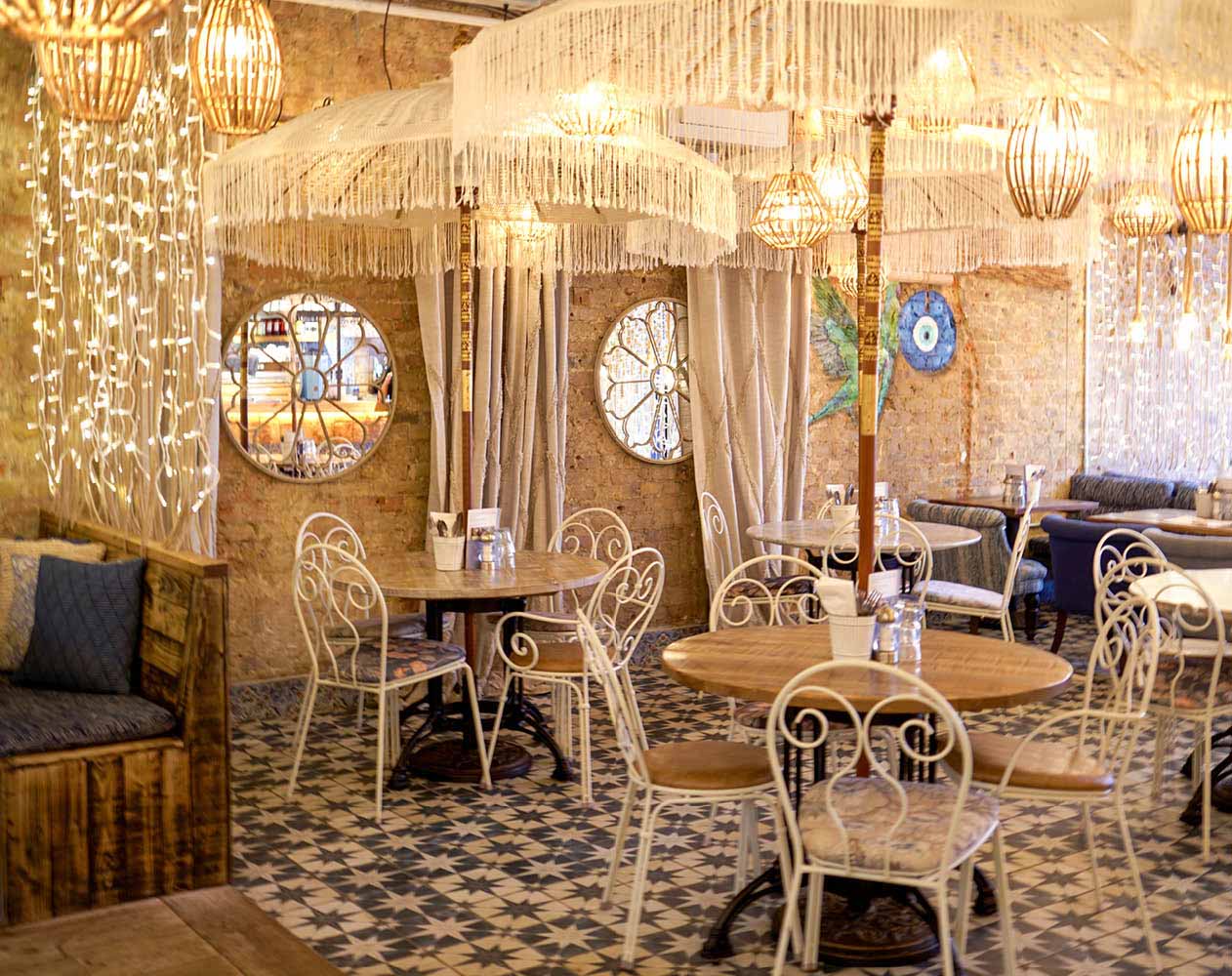 A cafe dining area with a vintage or eclectic style. The floor is covered in patterned tiles with a star motif. Several circular booths with light-colored, tufted upholstery and small round tables are arranged throughout the space. Ornate, white metal chairs with decorative backs surround the tables. The walls are exposed brick, with two round windows visible in the background. Numerous elaborate, fringed light fixtures hang from the ceiling, providing warm illumination. A wooden booth with a dark blue cushion is visible on the left. A portion of a dark blue sofa is visible on the right.